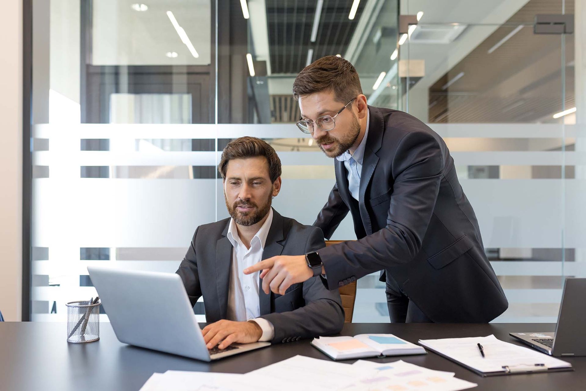 Two coworkers in an office reviewing a laptop, one standing and pointing at the screen.