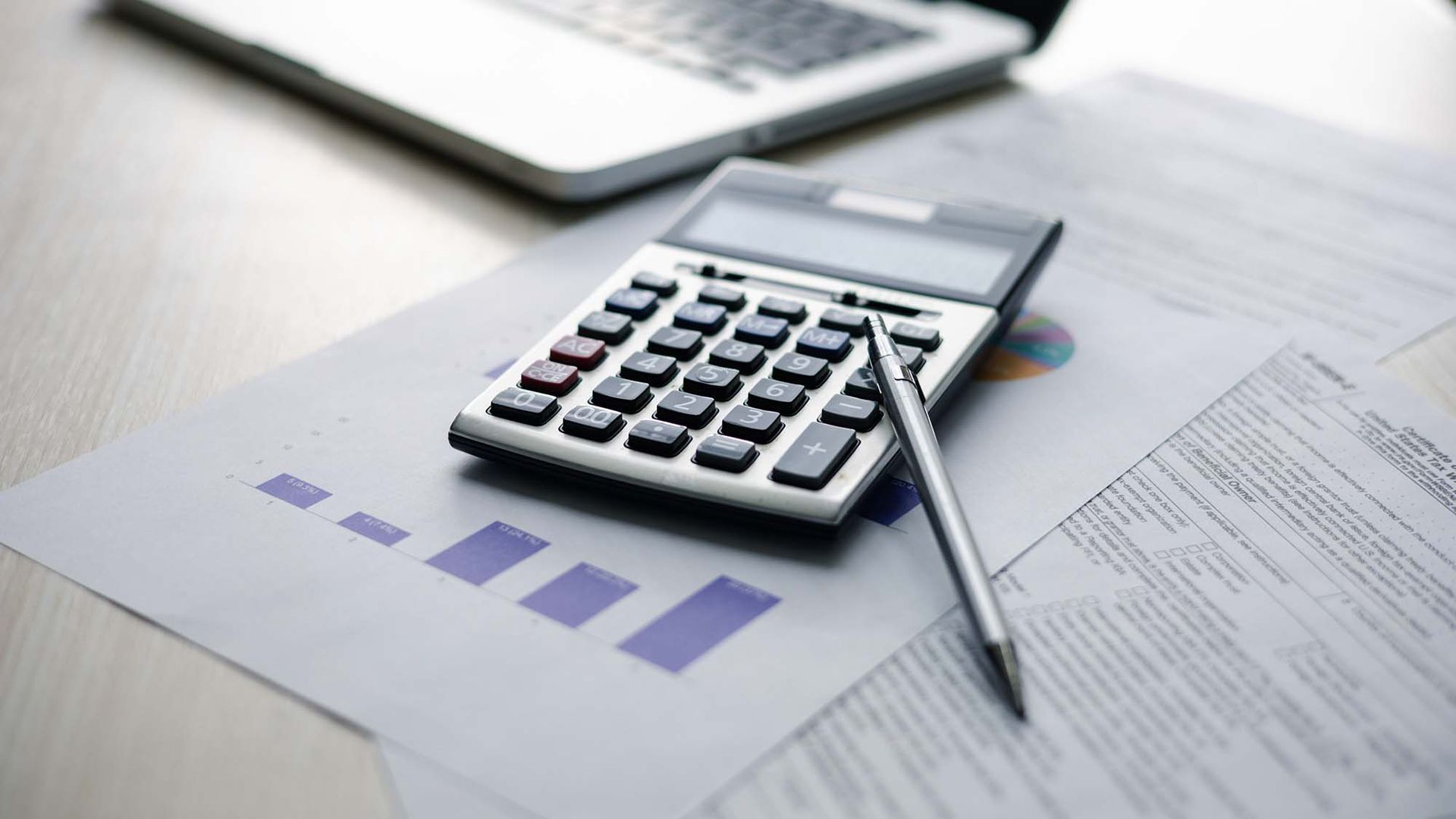 Calculator and pen on financial paperwork with laptop in the background, showing a budgeting or accounting scene