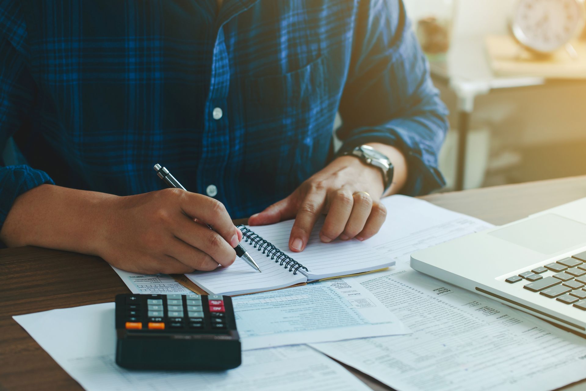 Person writing on paperwork beside a calculator and laptop at a desk