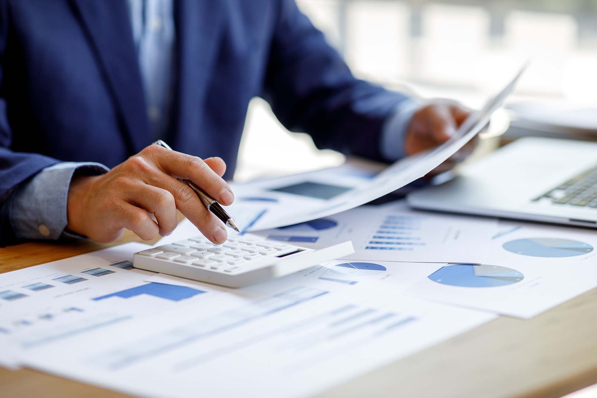Person analyzing printed charts with a calculator at a desk, with a laptop nearby.