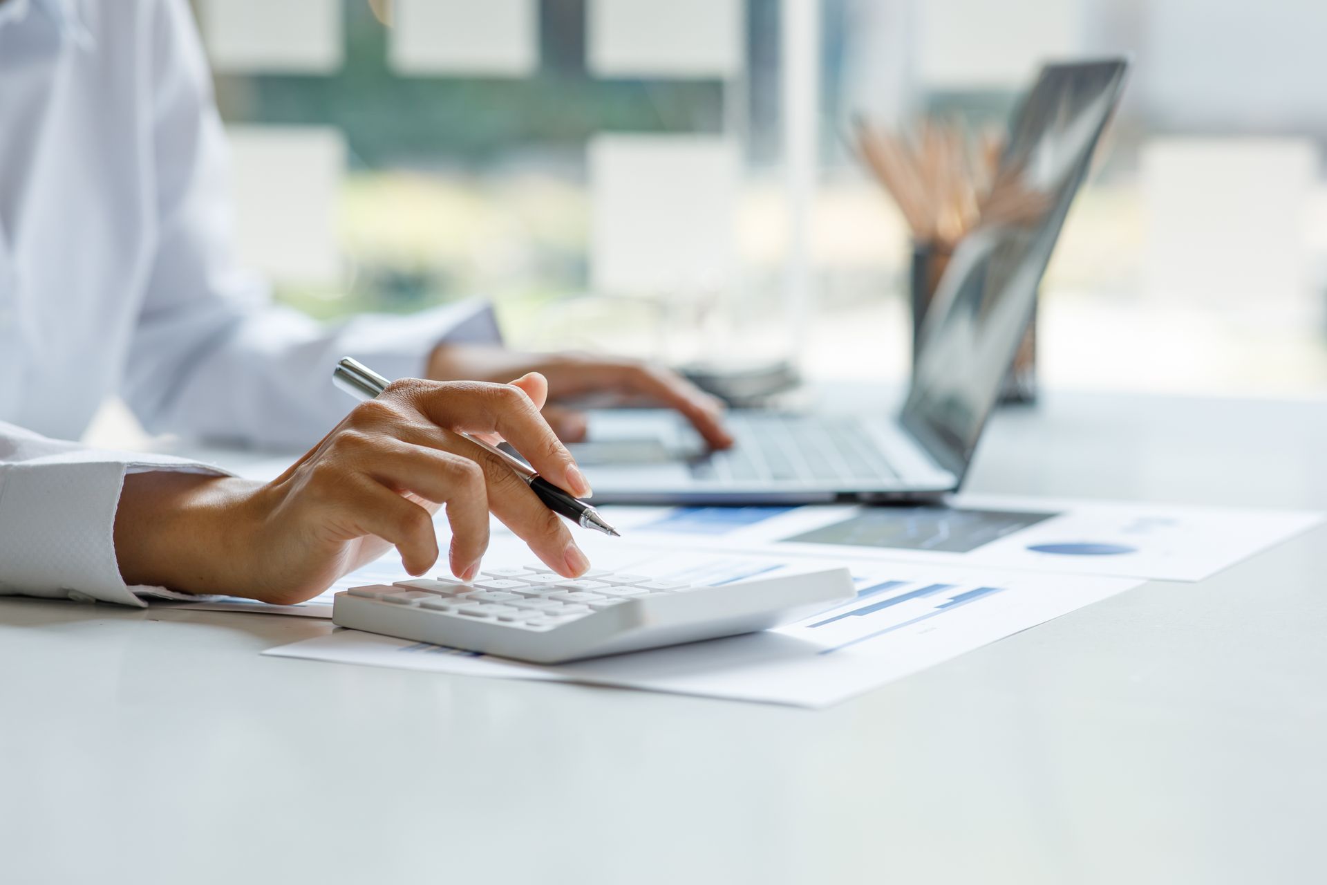 Hands writing on paper beside a laptop in a bright office setting