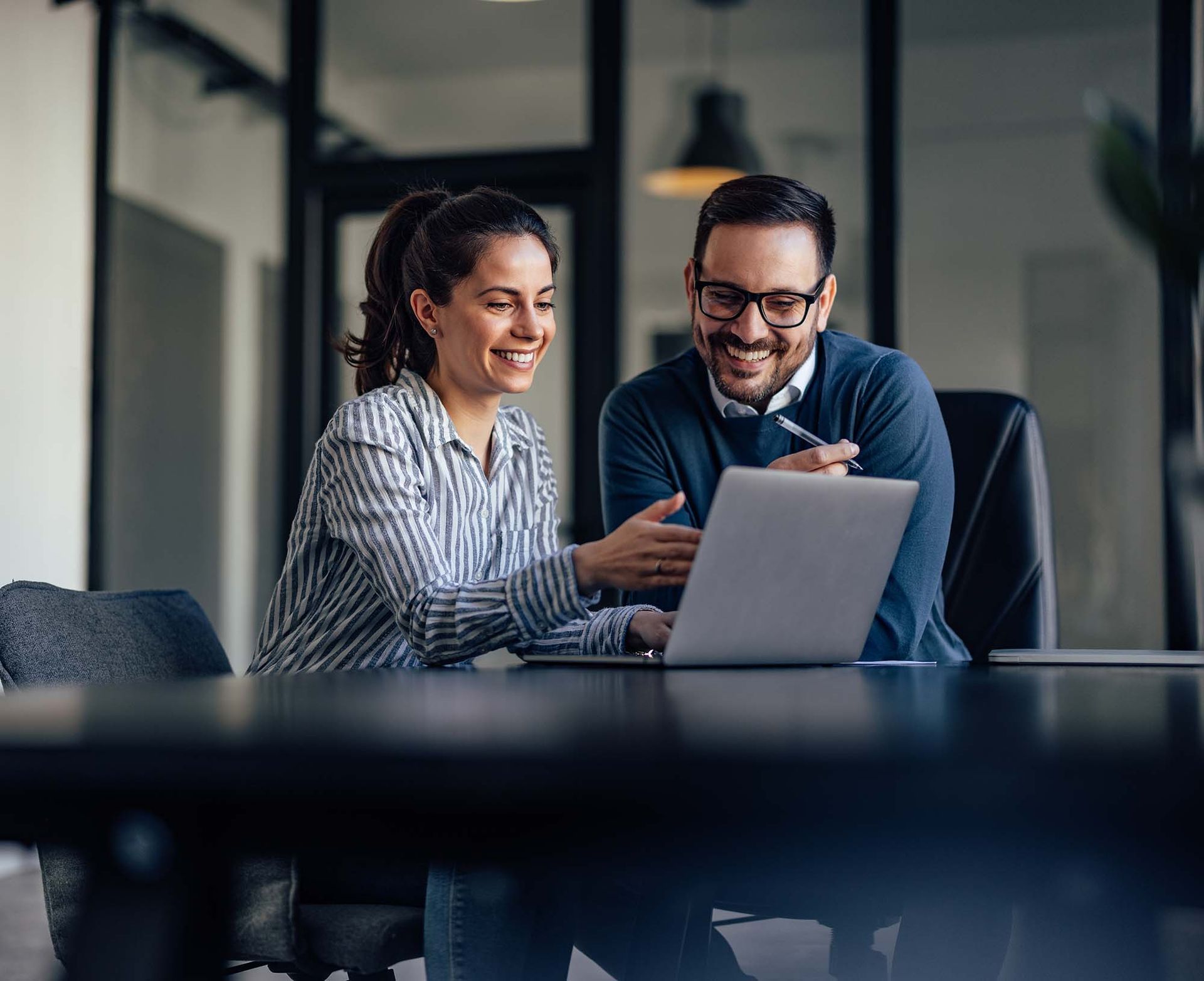 Two coworkers smiling and reviewing a laptop in a modern office