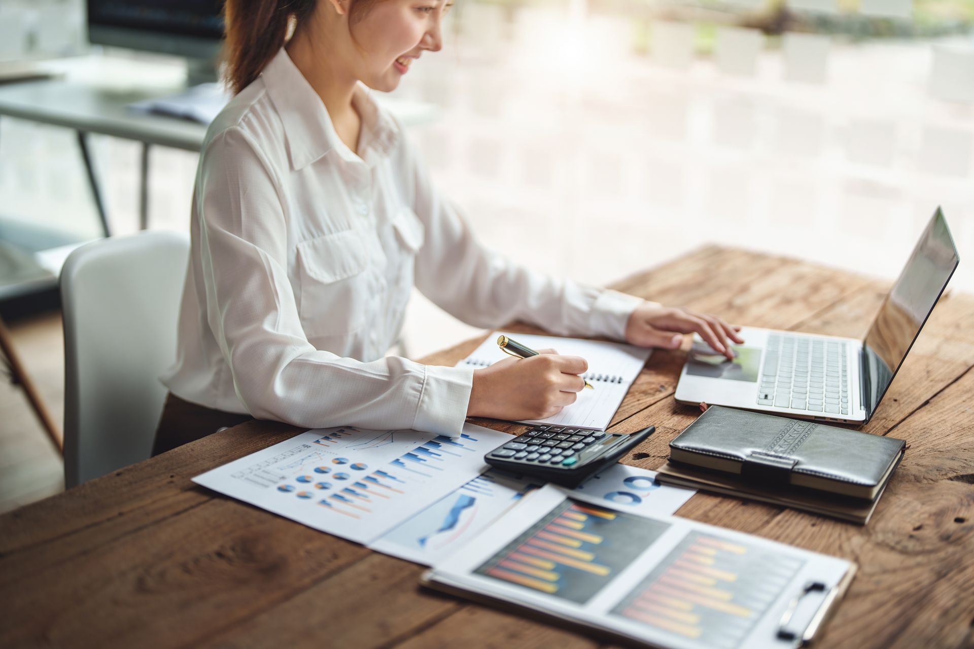 Person working at a desk with a laptop, calculator, and financial charts in a bright office