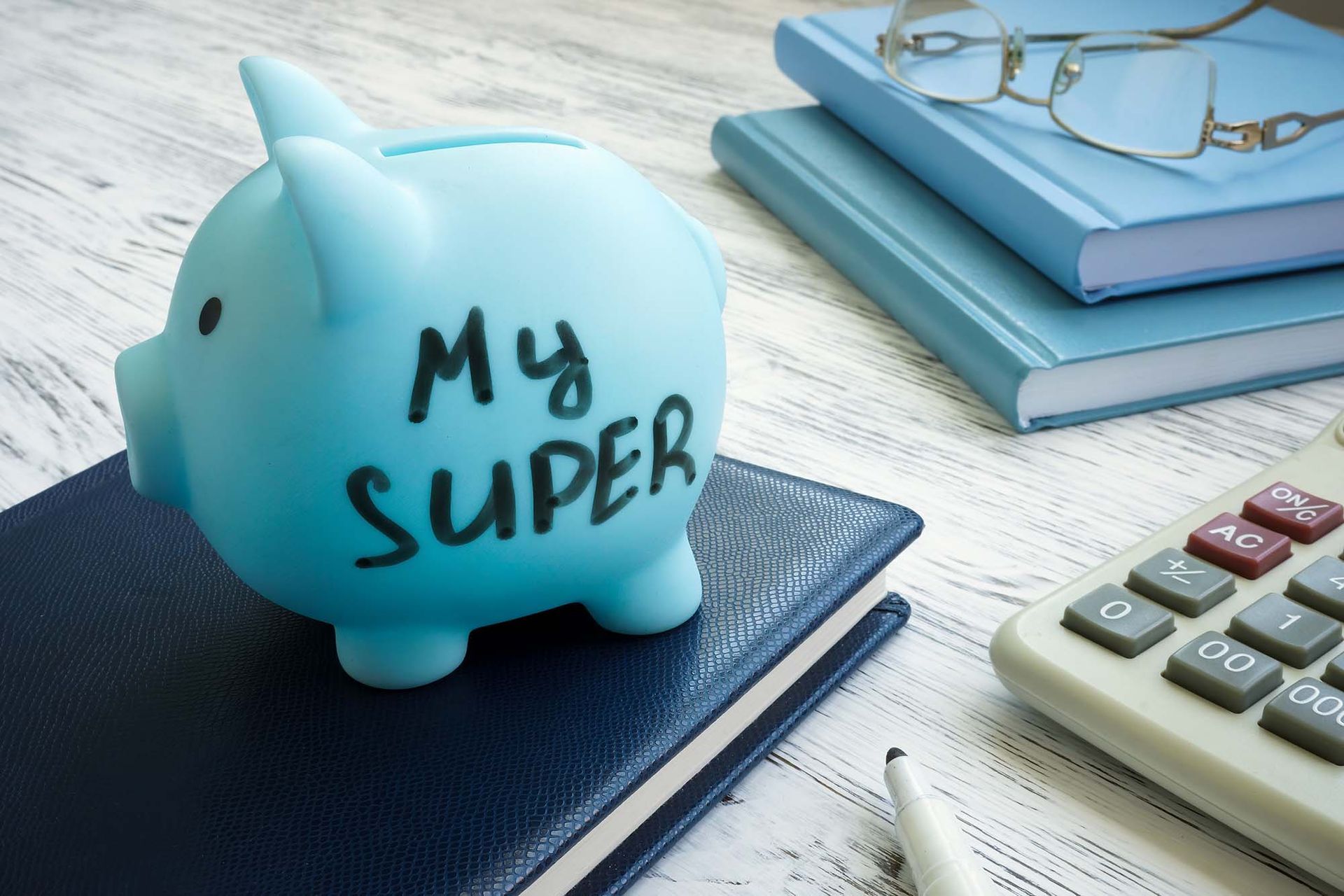 Light blue piggy bank labeled “My Super” on a desk with calculator and notebooks