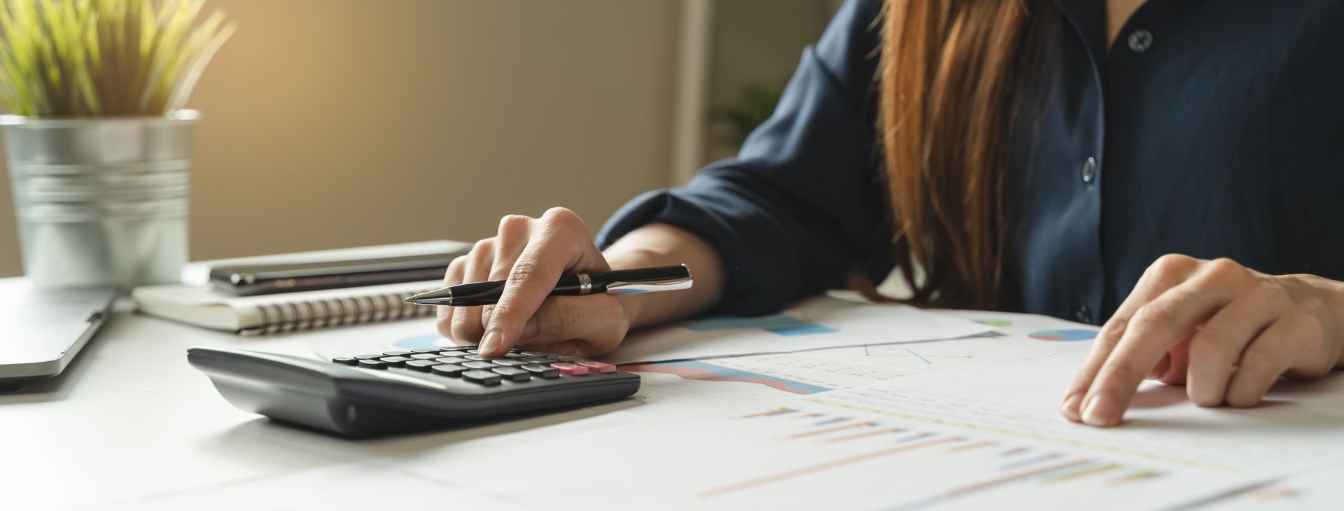 Person using a calculator at a desk with papers, laptop, and a small plant nearby