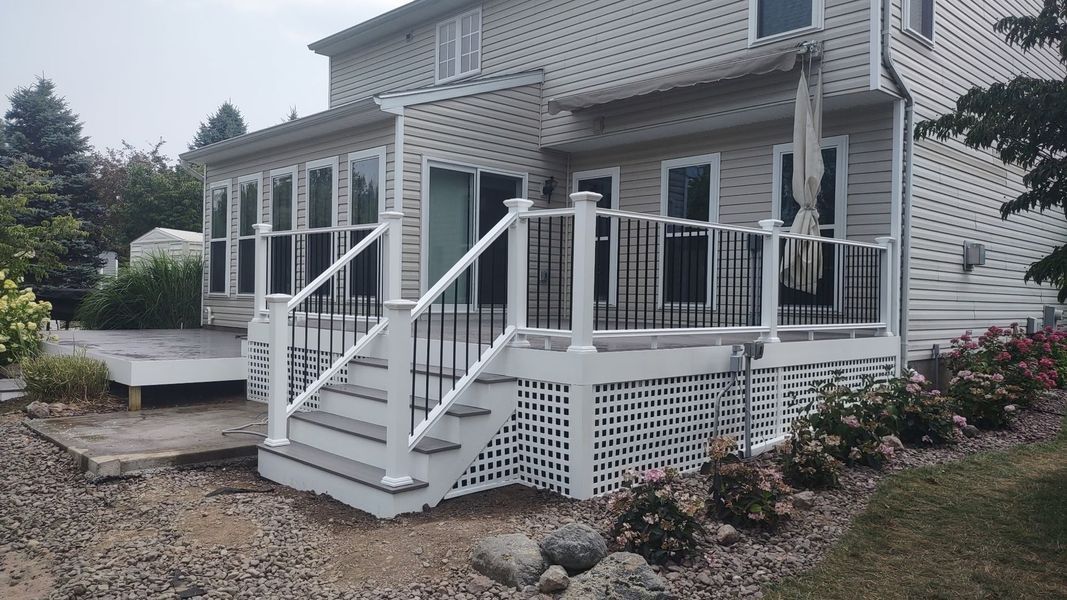 White house with wraparound porch and stairs, surrounded by landscaping and a stone patio.