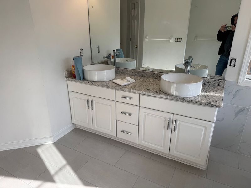 Bathroom vanity with double sinks, granite countertop, and white cabinets in a bright tiled room