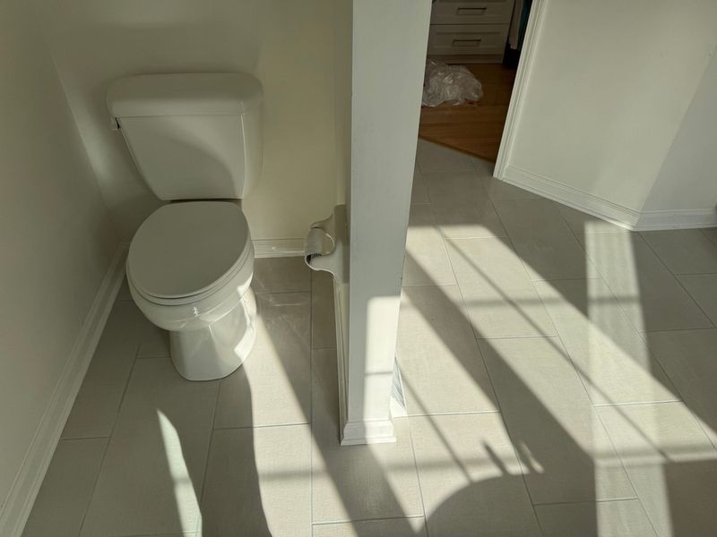Small bathroom with a white toilet, tiled floor, and sunlight casting shadows through the doorway.