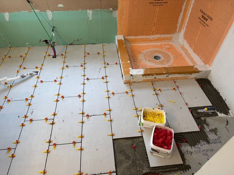 Partially tiled bathroom floor with orange wall tiles, gray mortar patches, and buckets of red and yellow tile adhesive