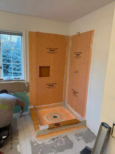 Bathroom shower under construction with orange waterproof backer board and a worker kneeling by the window.