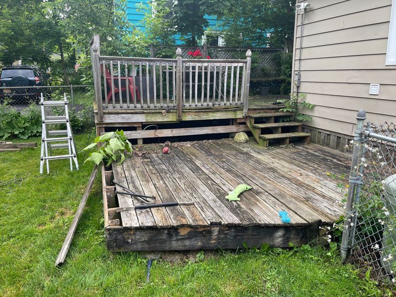 Weathered wooden deck with railing and ladder beside a house, with scattered debris and a small tree nearby