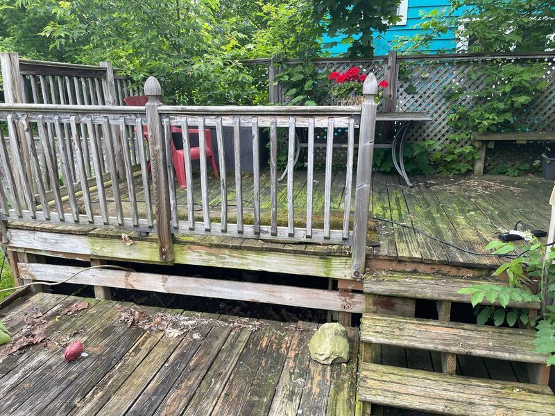 Weathered wooden deck with railing, steps, and patio furniture in a leafy backyard.