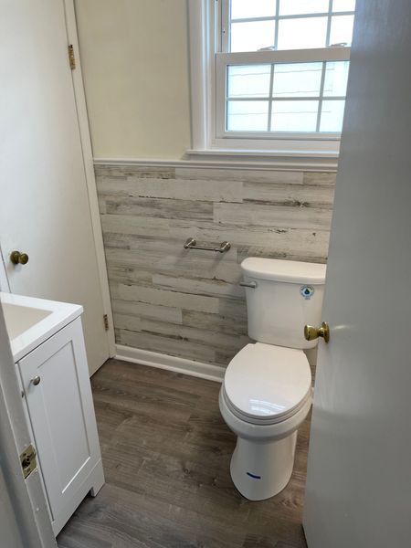 Small bathroom with white toilet, vanity, and gray wood-look wall paneling by a window.