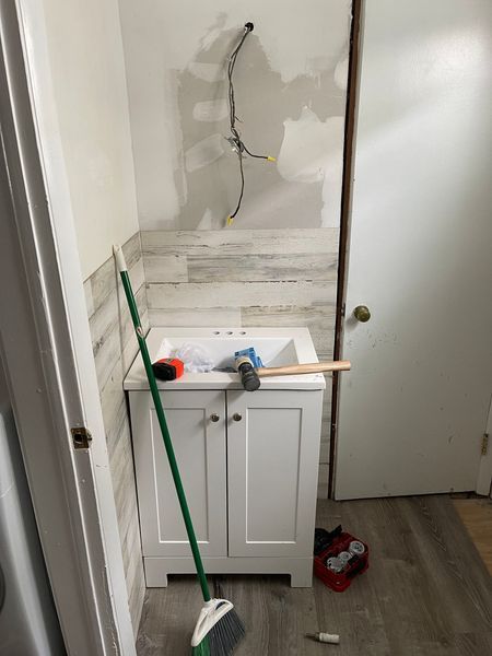 Damaged bathroom sink wall with crack, exposed pipes, and cleaning tools leaning against the vanity.