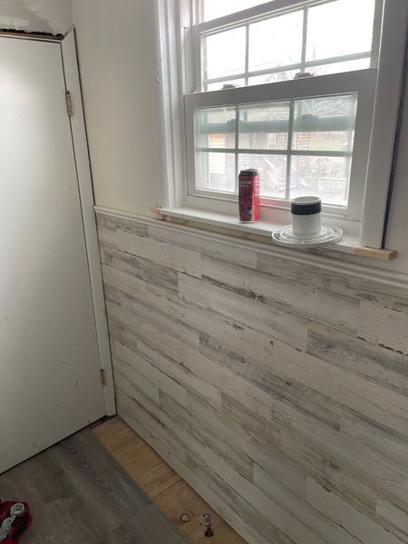 Small room with white wainscoting, window ledge, and a door; a red bottle and black container sit on the sill.