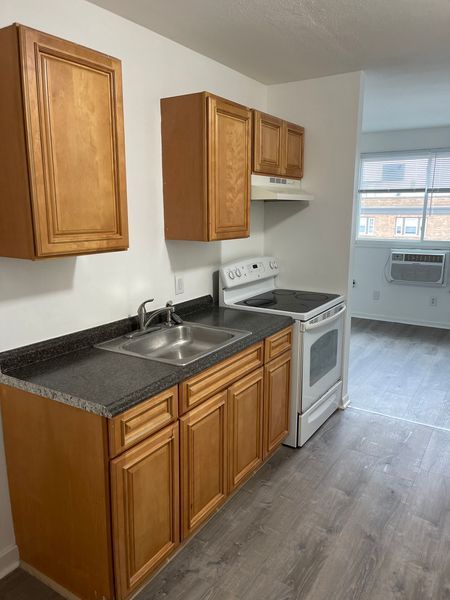 Small kitchen with wood cabinets, black countertops, white stove, and a window air conditioner in the next room