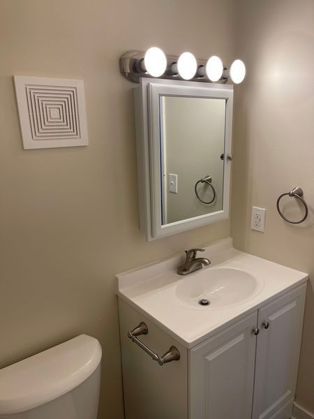 Small beige bathroom with sink, mirror, vanity lights, towel ring, and partial toilet visible.