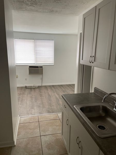 Small kitchen opening into a living room with tiled floor, gray cabinets, and a window air conditioner.