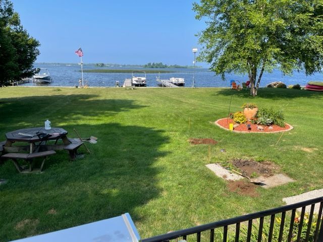 Lakeside lawn with picnic tables, flower bed, trees, and docked boats under a clear sky