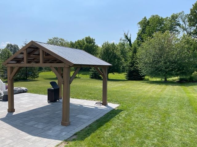 Wooden gazebo on a stone patio beside a green lawn under a clear blue sky