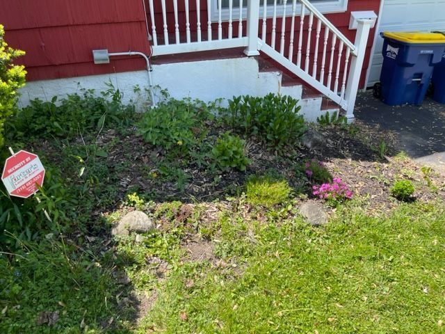 Front yard garden bed with green plants, a red house, white porch railing, and a security sign on the lawn.