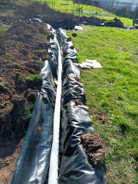 Black plastic-lined trench or irrigation ditch running through grassy soil outdoors.