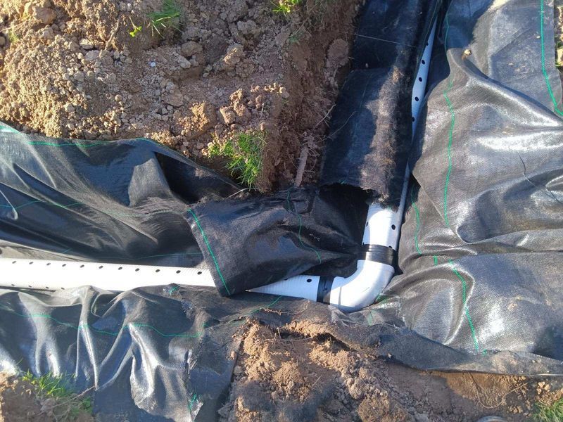 Black landscaping fabric around a white PVC pipe elbow in a dirt trench