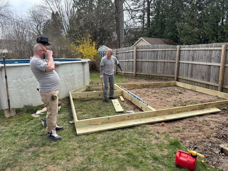 Two people assembling a raised garden bed in a backyard, with tools and lumber on the grass.