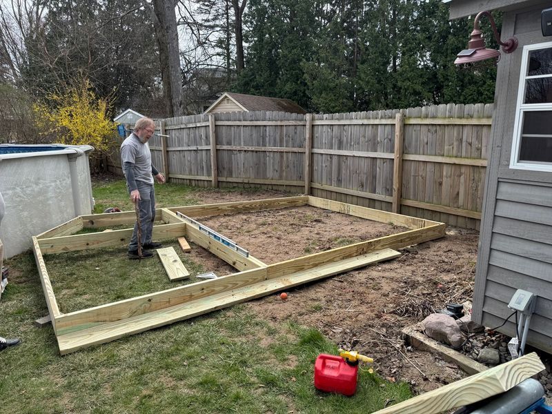 Person framing a small backyard deck beside a house, with lumber and tools on a fenced yard.