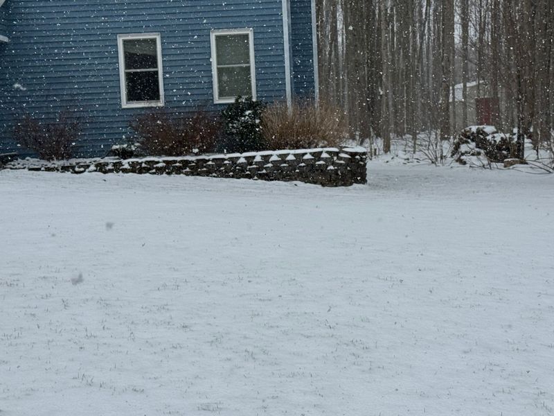Snow-covered yard beside a blue house with a stone retaining wall during snowfall