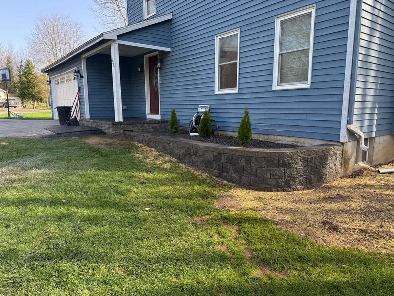 Blue house exterior with white trim, front porch, and small landscaped retaining wall beside a grassy yard