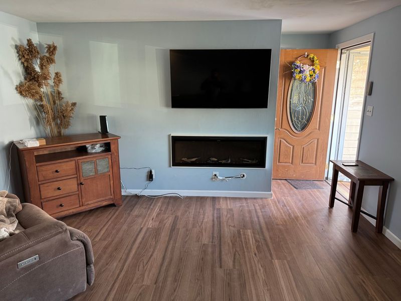Living room with wall-mounted TV, electric fireplace, wood floor, and front door with oval glass insert