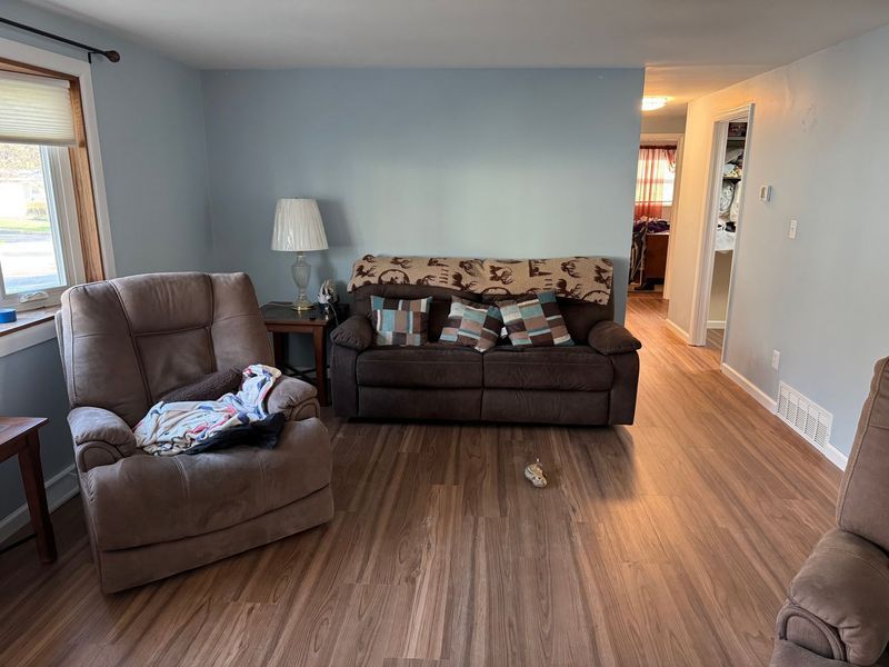 Living room with brown sofas, a floor lamp, and wood-look flooring; hallway visible in back.