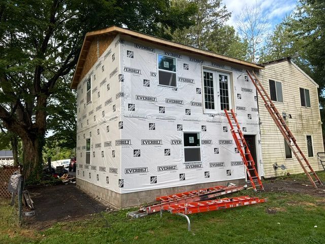 House under renovation with white siding wrap, ladders, and trees in the yard