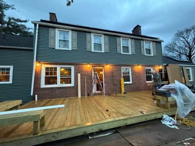 Back deck under construction at a gray two-story house, with a ladder and tools visible at dusk