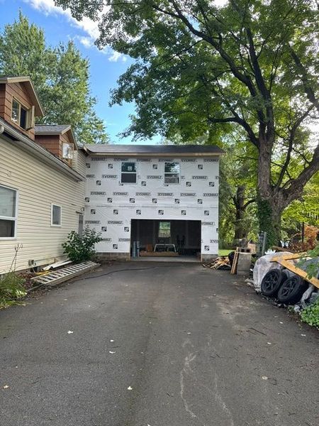 Driveway leading to a house under construction, with white siding, trees, and building materials on the right.