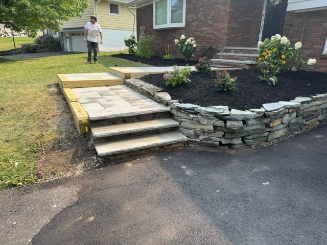 Front yard with stone steps and retaining wall beside a brick house; person standing near the walkway.