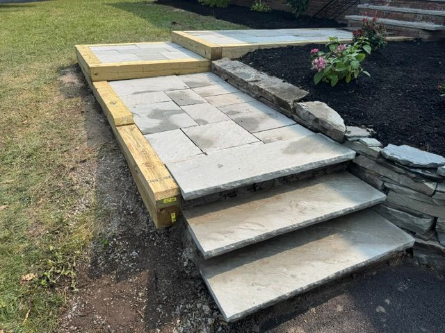 Stone front steps and walkway beside a garden bed, with one step cracked and uneven.