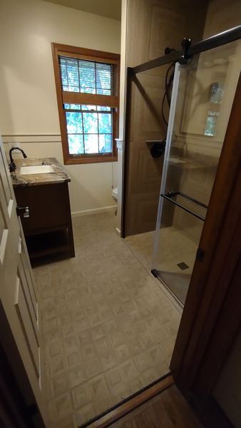 Small bathroom with vanity, window, and glass shower door on beige tile floor.