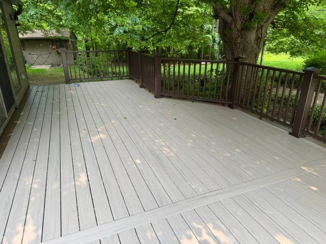 Wooden deck with brown railing under large green trees in a shaded backyard.