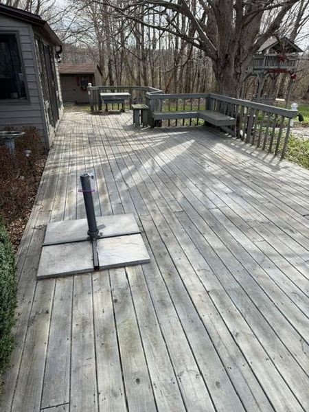 Wooden deck with railings, benches, and a tilted umbrella base in a wooded backyard.