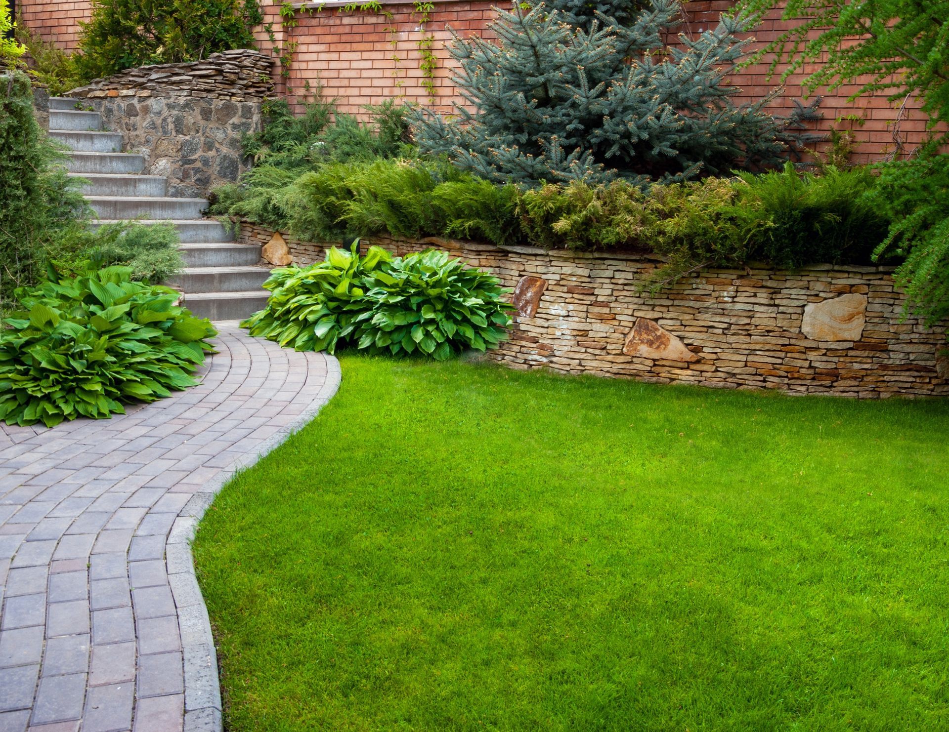 Curved stone path beside a green lawn and layered garden beds with shrubs, steps, and a brick wall