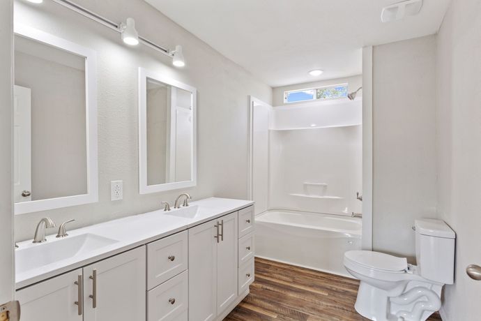 Bright modern bathroom with double vanity, large mirrors, tub, toilet, and wood-look floor.