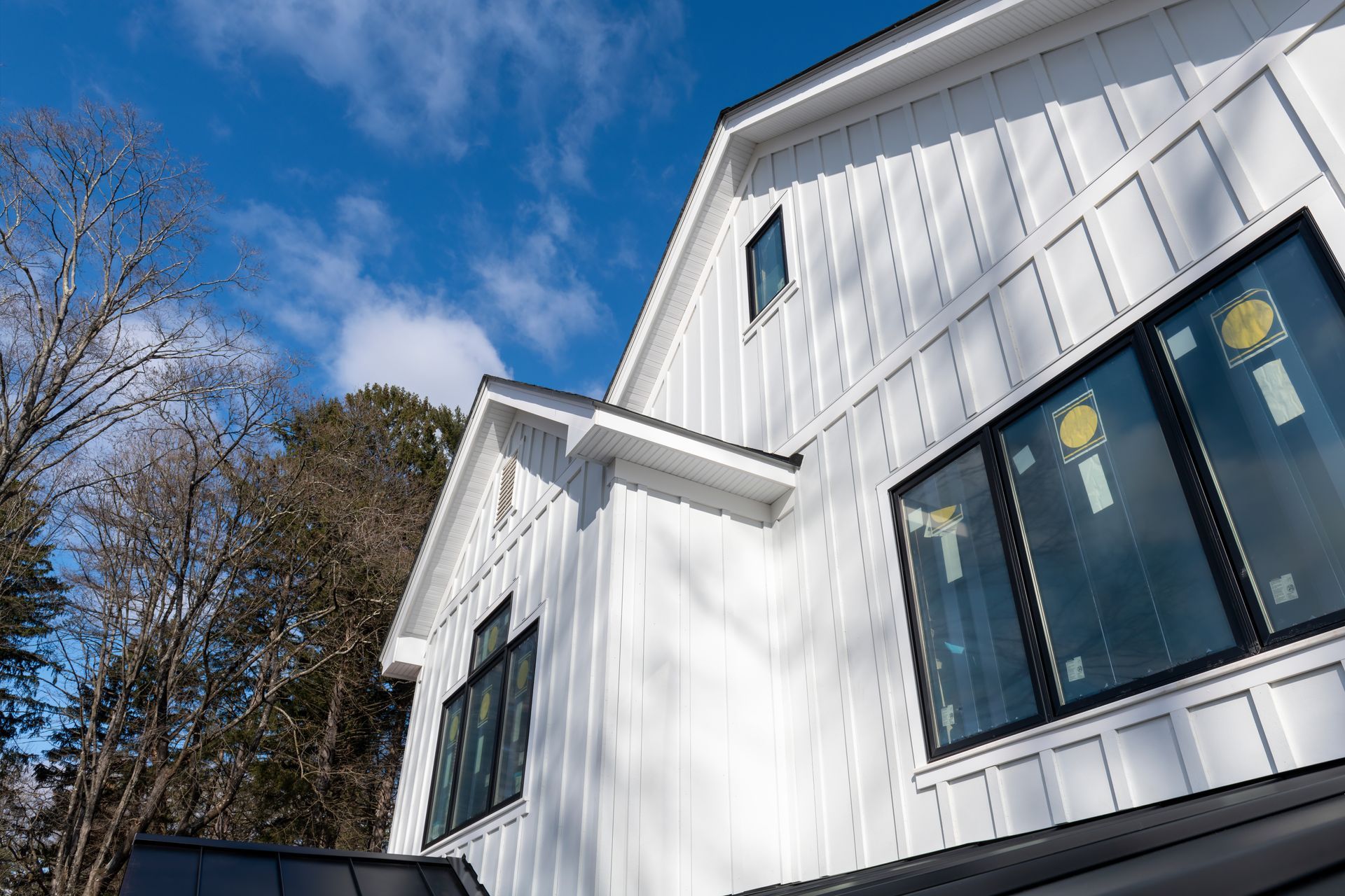 White house exterior with large windows and steep roof against a blue sky, framed by leafless trees