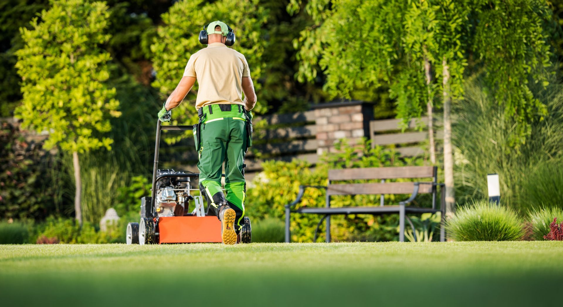 Person mowing a lawn in a green park, with a bench and trees in the background.