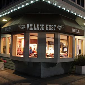 Village Host Pizzeria and Grill storefront at night, warmly lit with large windows and a corner entrance