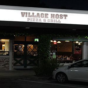 Village Host Pizza & Grill storefront at dusk, with dimly lit windows and a parked car in front