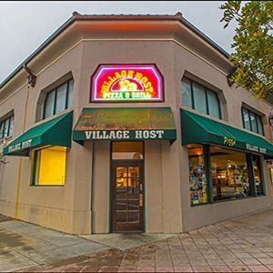 Corner storefront with green awnings and neon sign reading “Village Host” at dusk.