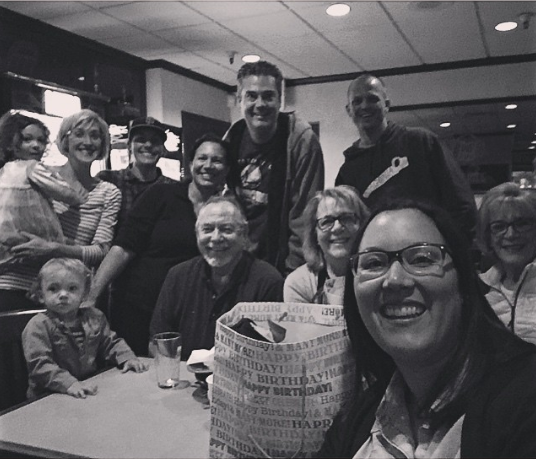 Group of people smiling around a table in a restaurant, with one person taking a selfie in front.