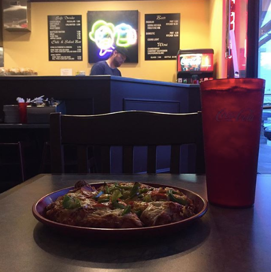 Restaurant table with pizza and red drink in foreground, booths and neon sign glow in the background